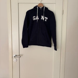 Marinblå hoodie från GANT med dragkedja - Snygg marinblå hoodie från GANT med vit logga på bröstet och vita snören i huvan. Tröjan har dragkedja framtill, två fickor och är tillverkad i mjuk bomull. Perfekt för chill dagar eller när du vill ha en klassisk streetstyle-look.