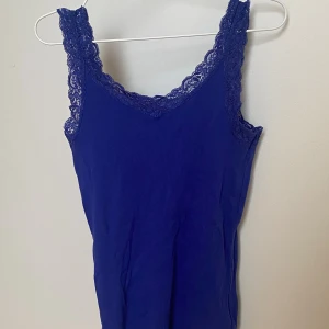 Blå linnetopp med spetskant och lace - Blå linnetopp med v-ringning och dekorativ spetskant längs axelbanden och halsringningen. Går att fråga om prissänkning. Tvättas innan den skickas