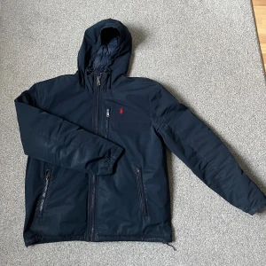 Marinblå vindjacka från Polo Ralph Lauren - Snygg marinblå vindjacka från Polo Ralph Lauren med huva, dragkedja framtill och två fickor med dragkedja. Jackan har röd broderad logga på bröstet och justerbar huva. Perfekt för blåsiga dagar och stilren look. Jackan saknar ens dragflärpen på ena fickan framtill. Annar också en liten fläck framtill på bröstet som syns i perfekt dagsljus! Pris kan diskuteras vid snabbaffär! Hör av dig vid fler frågor och funderingar! 