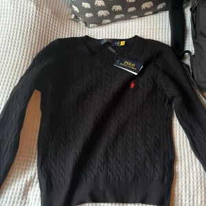 Svart kabelstickad tröja Polo Ralph Lauren  - (ALDRIG ANVÄND)Svart kabelstickad tröja från Polo Ralph Lauren med klassisk röd logga broderad på bröstet. Tröjan har rund halsringning, lång ärm och ribbade muddar. Perfekt för dig som gillar stilren och tidlös design. 