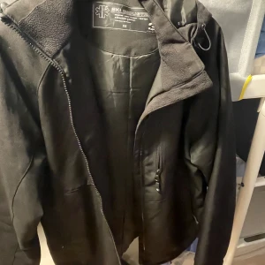 Svart vindjacka från Ski Industries XS - Svart vindjacka från Ski Industries i storlek XS. Jackan har huva med dragsko, flera fickor med dragkedja och mjukt fleecefoder på insidan av kragen. Perfekt för kyliga dagar och outdoor-aktiviteter.