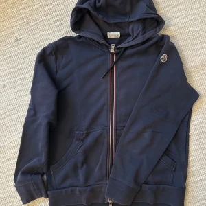 Mörkblå hoodie från Moncler - Snygg mörkblå hoodie från Moncler med dragkedja och huva. Tröjan har fickor framtill, Moncler-logga på ärmen och coola ränder längs dragkedjan. Tillverkad i 100% bomull för en skön och avslappnad känsla.