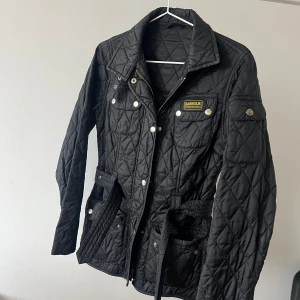 Svart quiltad jacka från Barbour - Quiltad svart jacka från Barbour International med snygga detaljer som guldiga tryckknappar, dragkedja med Barbour-ring och bälte i midjan. Klassisk krage, flera fickor och Barbour-logga på bröstet. Perfekt för höst och vinter.