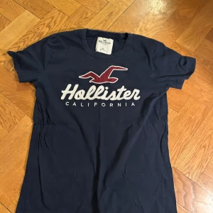 Mörkblå Hollister t-shirt med tryck - Snygg mörkblå t-shirt från Hollister med klassiskt logotryck och röd fågel på bröstet. T-shirten är i bomull och har rund hals och korta ärmar. Perfekt för en avslappnad och cool stil.