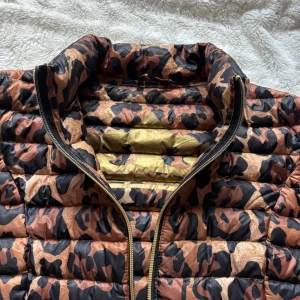 Leopardmönstrad pufferjacka med dragkedja - Snygg pufferjacka med leopardmönster i bruna och svarta toner. Jackan har guldig dragkedja framtill och på fickorna, hög krage och figurnära passform. Perfekt för dig som vill sticka ut med en cool look. Jackan är light weight och får plats i en liten tillhörande påse 