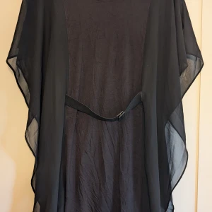 Svart kortklänning med bälte från H&M - Svart kortklänning från H&M i medium med transparenta, flödande ärmar och ett smalt bälte i midjan. Klänningen är kroppsnära men armarna ger den en lätt och luftig intryck. Är tillverkad i polyester. Perfekt för dig som gillar stilrena men piffiga plagg. Klänningen är enbart använd vid ett tillfälle. Hittar inget att anmärka på, möjligtvis att den behöver strykas 😅