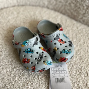 Grå Crocs med bilar, barnstorlek C7 - Säljer ett par ljusgrå Crocs med coolt bil-mönster i rött, blått och svart. Skorna har klassisk Crocs-design med rem bak och ventilerande hål framtill. Helt ny med lappar storlek 23-24