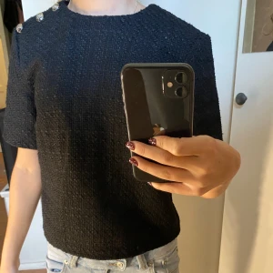 Svart kortärmad blus från H&M - Säljer en svart, strukturerad topp från H&M i storlek XS. Modellen är boxig med korta ärmar och rund halsringning. Tre dekorativa knappar i silver på ena axeln ger en snygg detalj. Toppen har dragkedja i ryggen och är gjord i ett grovt, vävt material.