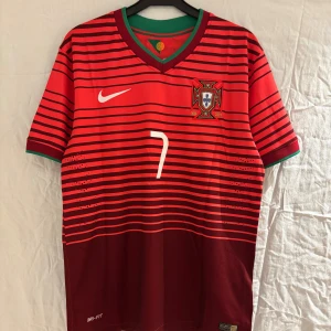 Portugal Ronaldo 7 fotbollströja Nike M - Portugal landslagströja från Nike med Ronaldo 7-tryck. Röd och vinröd med svarta horisontella ränder, gröna detaljer vid krage och ärmslut. Broderat landslagsmärke, Dri-FIT-material för bra andningsförmåga. Retro 2014-modell.