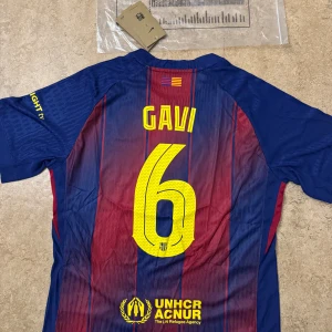 FC Barcelona Gavi matchtröja Nike - Barça matchtröja med Gavi 6 på ryggen, klassiska blå och röda ränder, gult Nike-märke och Spotify-logga. Broderat klubbmärke på bröstet. Tillverkad i lätt, ventilerande polyester – perfekt för fotbollsplanen eller läktaren.