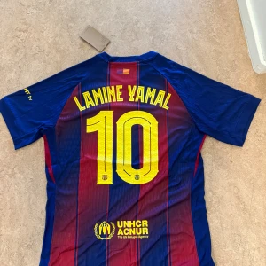 FC Barcelona Lamine Yamal tröja Nike L - Säljer en officiell FC Barcelona matchtröja från Nike med Lamine Yamal och nummer 10 på ryggen. Tröjan är blå och röd med gula detaljer, randigt mönster och klubbmärke på bröstet. Tillverkad i lätt och ventilerande Dri-Fit material.