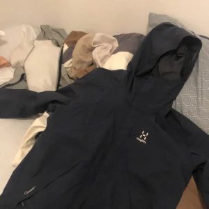 Svart vindjacka från Haglöfs - Säljer en svart vindjacka från Haglöfs med huva och diskret logga på bröstet. Jackan har praktiska fickor och är tillverkad i slitstark syntet. Perfekt för blåsiga dagar och outdoor-äventyr. Snygg och funktionell design.