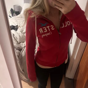 Röd hoodie från Hollister med huva - Säljer en röd hoodie från Hollister med vit text och blå logga på bröstet. Tröjan har dragkedja, snörning vid huvan och mysigt foder i huvan. Jätte skön och i super skick!