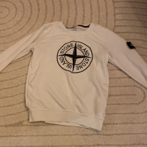 Vit Stone Island sweatshirt S - Vit sweatshirt från Stone Island i storlek S med stort svart logotryck på bröstet och klassisk patch på vänster ärm. Tröjan har rund hals och långa ärmar, tillverkad i mjuk bomull. Perfekt för dig som gillar streetwear och vill sticka ut med en ikonisk look.