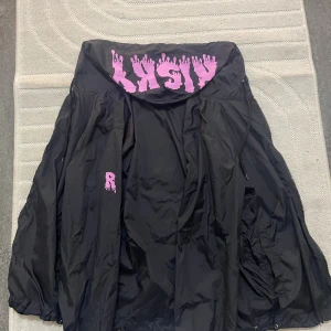 Svart vindjacka från Risky XS - Svart vindjacka från Risky med stor huva och coolt rosa tryck på lovan och armen. Jackan har dragsko i huvan och är tillverkad i lätt polyester. Perfekt för dig som vill sticka ut med streetstyle-vibe.