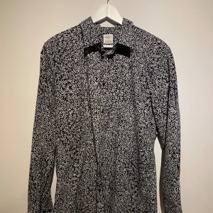 Svartvit mönstrad skjorta Morris & Co x HM - Snygg svart skjorta från Morris & Co x H&M med vitt bladmönster över hela tyget. Skjortan har klassisk krage, långa ärmar och knäppning framtill. Bomull! 