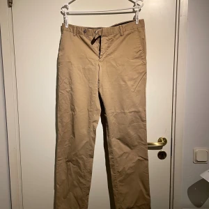 Beiga chinos från Oscar Jacobson - Snygga beiga chinos från Oscar Jacobson med klassisk rak passform. Byxorna har bälteshällor, knappstängning och fickor både fram och bak med knappdetaljer. 