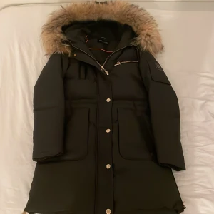 Svart parkas från Hollies med fuskpäls - Svart lång parkas från Hollies med stor huva och fluffig fuskpäls. Jackan har guldiga dragkedjor, tryckknappar och flera fickor, inklusive en på ärmen med Hollies-logga. Perfekt för kalla vinterdagar och riktigt snygg streetstil.