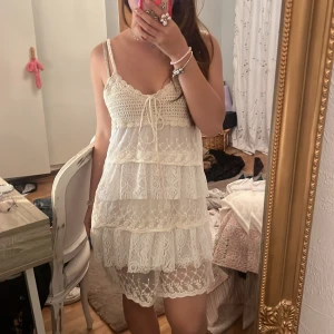 Vit spetsklänning med snörning - Supersöt vit kortklänning med tunna axelband och omlott snörning framtill. Klänningen har flera lager med spets och virkade detaljer upptill, vilket ger en romantisk och bohemisk vibe. Perfekt för sommarens alla tillfällen och lätt att styla med smycken.