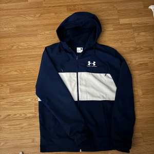 Blå och vit vindjacka Under Armour XS men funkar även som S  - Snygg vindjacka från Under Armour i marinblått med vit panel över bröstet och logga på vänster sida. Jackan har huva med snörning, hel dragkedja och meshfoder. Perfekt för dig som gillar sportig stil och vill ha något lätt och funktionellt.
