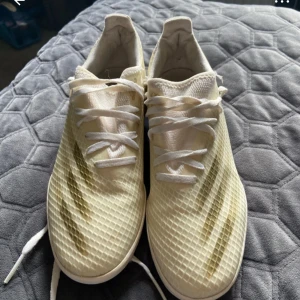 Vita fotbollsskor från Adidas - Säljer ett par vita fotbollsskor från Adidas med klassiska tre ränder i guld på sidan. Skorna har snörning och ovandel i syntetmaterial med ett mönstrat nätliknande yta. Perfekta för dig som vill sticka ut på planen!