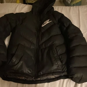 Svart pufferjacka från Nike - Svart pufferjacka från Nike med stor vit logga på bröstet. Jackan har dragkedja framtill, två sidofickor och är fylld med dun och polyester. Den har en klassisk pufferlook med breda, vadderade sektioner och långärmade ärmar. Perfekt för kalla dagar.Lite trög dragkedja ,för övrigt i fint använt skick.