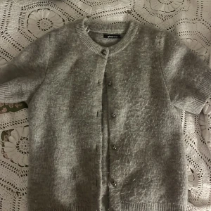 Grå stickad kofta från Gina Tricot - Mysig grå stickad kofta från Gina Tricot i storlek S. Koftan har korta ärmar, rund hals och knappar framtill. Perfekt för lager-på-lager och har en fluffig, mjuk känsla. Lite nopprig, men går enkelt att ta bort med maskin! ❤️