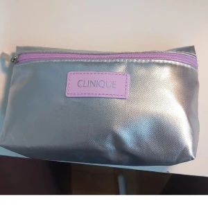 Silver necessär från Clinique med hudvård - Snygg och stilren necessär från Clinique i metallic silver med ljust lila dragkedja och matchande lila logotyp. Perfekt storlek för smink eller hudvård, med mjuk och glansig yta i syntetmaterial. Enkel att ta med i väskan och håller dina produkter organiserade. Inuti finns det en 60ml Clarifying Lotion, en 15ml 100H Auto-Replenishing Hydrator, en 15ml Cleansing Balm, en 10ml Dark Spot Correcter/Interrupter, och två 5ml Wrinkle Correcting Eye Cream/Serum.
