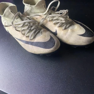 Nike Mercurial fotbollsskor beige - Säljer ett par Nike Mercurial fotbollsskor i beige med svarta detaljer och stor Swoosh-logga. Skorna har snörning, stickad ovandel och plös, samt dobbar för gräsplan. Lätta och smidiga med en modern, strumpliknande passform.