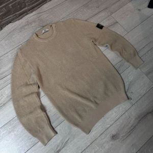 Beige stickad tröja från Stone Island - Snygg beige stickad tröja från Stone Island med klassisk rund hals och ribbade muddar. Tröjan har den ikoniska Stone Island-patchen på vänster ärm och är gjord i ett grovstickat material som ger en cool och avslappnad vibe. PASSAR ÄVEN M😁😁 och tröjan är aldrig använd ✌️👍