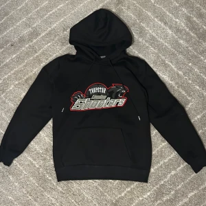Trapstar svart hoodie med broderi - Svart hoodie från Trapstar med stort broderat 'London Shooters'-motiv på bröstet i grått, vitt och rött. Tröjan har huva med snörning, ribbade muddar och klassisk känguruficka. Perfekt för dig som gillar streetwear och vill sticka ut. (Finns med byxor också)