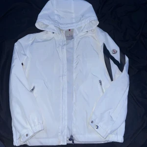 Vit vindjacka från Moncler med huva - Snygg vit vindjacka från Moncler med huva och dragkedja framtill. Jackan har svarta detaljer på ena ärmen och Moncler-logga på axeln. Utrustad med två fickor med dragkedja och elastiska muddar vid ärmslut och nederkant. Perfekt för dig som gillar stilrena och sportiga plagg.