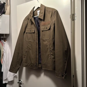 Olivgrön shirt jacka från Zara - Snygg olivgrön overshirt från Zara med klassisk krage, två stora bröstfickor med knappar och diskreta sidofickor. Jackan har dragkedja framtill och knappar vid ärmslut. Perfekt för dig som gillar stilren och enkel design.