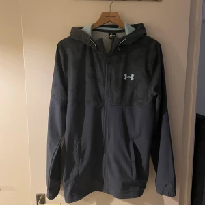 Mörkblå vindjacka från Under Armour - Snygg mörkblå vindjacka från Under Armour med camo-mönstrad övre del och ljusblå detaljer i huvan. Jackan har dragkedja framtill, två sidofickor och justerbar huva med snörning. Perfekt för dig som gillar sportig stil och funktion.