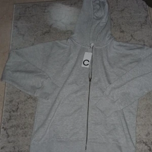 Grå hoodie med dragkedja från Cubus - Snygg grå hoodie från Cubus i storlek 158/164. Tröjan har huva, hel dragkedja framtill och två stora fickor. Tillverkad i mjuk bomullsmix som är skön att ha på sig. Perfekt för chill dagar och enkel att matcha med jeans eller joggers.