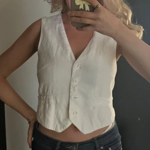 Vit linneväst från Pull&Bear - Snygg vit väst från Pull&Bear i linne med klassisk v-ringning och knappar framtill. Västen har två fejkade fickor och en figurnära passform som ger en clean och trendig look. Perfekt att styla över en t-shirt eller skjorta för en fräsch vibe.