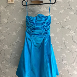 Blå satin klänning med snörning - Supersnygg blå klänning i glansigt satinmaterial med smala axelband och draperad överdel. Klänningen har superfin snörning i ryggen. Modellen är kort och köpt i nyskick men aldrig fått användning för.
