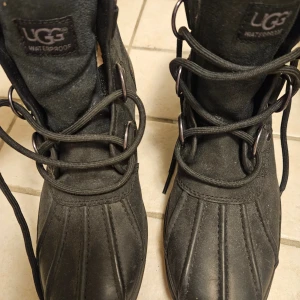 Helt nya Svarta vinterboots från UGG - Svarta UGG boots med snörning och vattentätt yttermaterial. Insidan är fodrad med mjuk päls för extra värme. Grov sula med UGG-logga undertill, perfekt för kalla dagar. Klassisk rund tå och robust design i skinn och syntet.