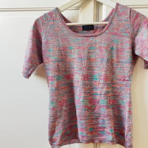 Glittrig flerfärgad topp från Sariia - Säljer en glittrig stickad topp från Sariia med korta ärmar och rund hals. Färgerna är en mix av rosa, turkos, lila och silver med ett melerat mönster. Materialet är mjukt och har en skimrande finish som syns tydligt i ljuset. Perfekt för dig som gillar färg och glitter!