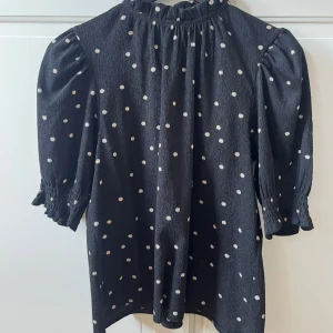 Svart prickig klänning Holly & Whyte - Supersöt svart klänning med vita prickar från Holly & Whyte by Lindex. Klänningen har puffiga trekvartsärmar med smock vid ärmslut, hög hals med liten volang och knäppning bak i nacken. Perfekt för dig som gillar lekfulla detaljer och klassiskt mönster.