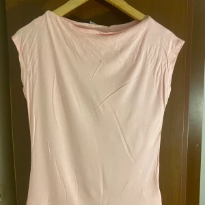 Ljusrosa T-shirt från Livia - Supersöt ljusrosa T-shirt från Livia i storlek S. Modellen är enkel och rak med ärmlös design och rundad hals. Materialet känns mjukt och har en lätt glans, perfekt för dig som gillar minimalistisk stil och ljusa färger.