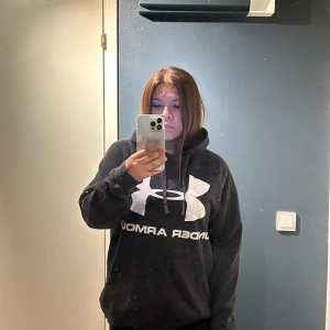 Svart Under Armour hoodie XL - Svart hoodie från Under Armour i storlek XL med stor vit logga och text på bröstet. Tröjan har huva med snörning och en rymlig magficka. Mjuk insida och ribbade muddar vid ärmslut och nederkant. 