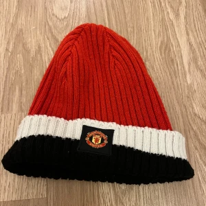 Manchester United röd mössa - Ribbstickad mössa i rött med bred vit och svart kant, prydd med Manchester Uniteds emblem framtill. Tillverkad i 100% akryl, perfekt för dig som vill visa ditt lagstöd även när det är kallt ute.