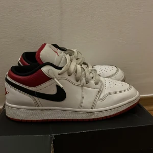 Nike Air Jordan 1 Low Chicago - Nike Air Jordan 1 Low i klassiska Chicago-färger. Skorna har vit läderbas, svarta swoosh-detaljer och röd häl samt yttersula. Röd Jumpman-logga på plösen och Air Jordan Wings broderat bak. Lågt skaft och snörning framtill. Perfekta för dig som gillar streetwear och sneakerskultur.