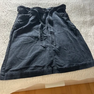 Svart manchesterkjol från H&M med knyt - Svart manchesterkjol från H&M i storlek 46. Kjolen har knytband i midjan, knappar vid fickorna och en rak, enkel design. Perfekt för dig som gillar en klassisk och avslappnad stil med lite extra struktur tack vare manchestertyget.