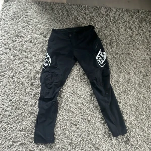 Troy Lee Designs Sprint V.28 byxor svart - Svarta Troy Lee Designs Sprint V.28 byxor med vita loggor på låren. Byxorna har smal passform, perforerade paneler för ventilation och är tillverkade i slitstarkt syntetmaterial. Perfekta för cykling eller enduro  med sportig look och smarta detaljer.