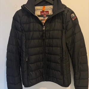 Svart light weight pufferjacka - Svart pufferjacka från Parajumpers med huva och quiltad design. Jackan har dragkedja framtill, två sidofickor med dragkedja och en snygg patch på ärmen. Lättviktig och perfekt för kyliga dagar.  Den är några år gammal och är väl använd men fortfarande hel. Köpt från kidsbeandstore och är i deras största storlek, kan jämföras med XS