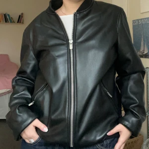 Svart skinn bomberjacka från Zara - Snygg svart bomberjacka från Zara i skinnimitation med ribbade muddar och silverfärgad dragkedja framtill. Jackan har två fickor med dragkedja och en stilren, loose passform, jag viker in kanterna längst ned för kortare jacka men det är givetvis valfritt 🥰