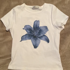 Vit t-shirt med blå blomma från FB Sister - Vit t-shirt från FB Sister i storlek S med en stor blå blomma tryckt på framsidan. Klassisk rund halsringning och korta ärmar. Perfekt för dig som gillar stilrena plagg med en twist. Materialet är mjuk bomull som känns skönt mot huden.