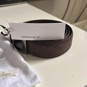 Brunt mockabälte från Onewear Sweden - Stilrent brunt bälte i mocka från Onewear Sweden. Bältet har en klassisk design med silverfärgat spänne och kommer i en snygg tygpåse. Perfekt accessoar för att lyfta din outfit med en touch av kvalitet och enkelhet. Fick den i mystery gift men var för liten